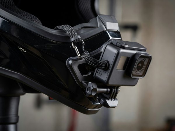 Action camera attached to motorcycle helmet chin bar using curved adhesive base and safety tether